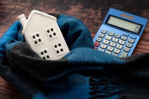 A small white model of a house wrapped in a scarf placed next to a calculator on a wooden surface.