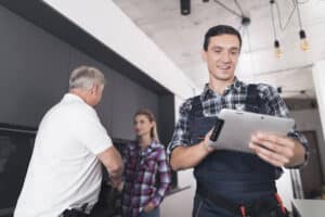 A woman shaking hands with a plumber while another plumber uses a tablet in a home.