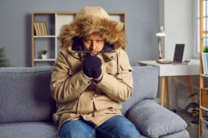 A man looking cold in a winter jacket and gloves as he sits on a couch in a home.